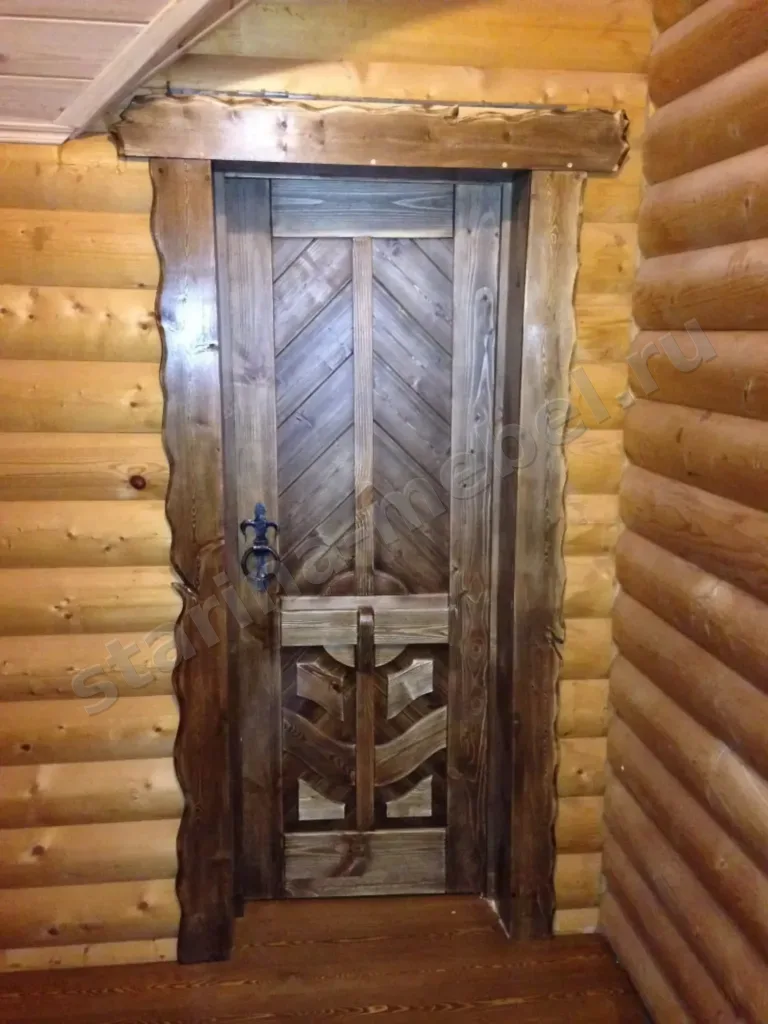 Тёмная дверь с выкладкой досок ёлочкой и резной нижней частью в бревенчатом доме